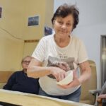 Starejša gospa posipa sladkor po razvaljanem testu na mizi, v ozadju sedi druga stanovalka in opazuje pripravo peciva.
