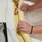 Prikaz rok, ki zvijajo razvaljano testo z nadevom v dolgo rolico za pripravo cimetovih rolic.