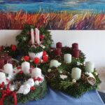 Na mizi je razstavljenih več adventnih venčkov iz zelenja, okrašenih s svečami različnih barv, suhim sadjem, storži in prazničnimi dodatki; v ozadju visi barvita slika pokrajine.