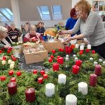 Starejše osebe sedijo za dolgo mizo, polno že izdelanih venčkov z belimi, rdečimi in vijoličnimi svečami; ena izmed udeleženk dodaja okraske iz škatle.