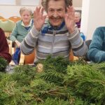 Starejša gospa v sivem puloverju in s pisanico okrog vratu veselo dviguje roke, medtem ko pred njo na mizi leži kup zelenih vejic za izdelavo adventnih venčkov.