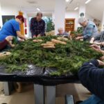 Skupina starejših oseb sedi okoli velike mize ter s pomočjo osebja oblikuje osnove za adventne venčke iz zelenih vej in slamnate podlage.