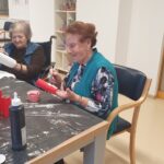 Dve stanovalke pri mizi barvata kartonske tulce z rdečo barvo in izdelujeta praznične dekoracije.