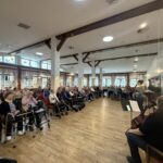 Dvorana doma, polna stanovalcev, ki sedijo v vrstah in poslušajo nastop; v ospredju nastopajoče z glasbenimi inštrumenti.