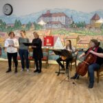 Skupina nastopajočih žensk poje ob spremljavi kitare in violončela pred poslikano steno z motivom mesta.