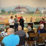 Nastopajoče pevke in glasbenici med nastopom, pred njimi pa sedi občinstvo stanovalcev na stolih in invalidskih vozičkih.