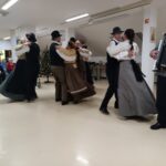 Folklorna skupina v tradicionalnih oblačilih pleše v dvorani doma starejših, ob strani harmonikar, v ozadju sedi občinstvo.