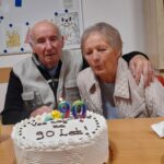 Starejši par sedi za mizo pred torto z napisom »Vse najboljše za 90 let« in številkama 90; gospa piha svečko, gospod jo objema okoli ramen.