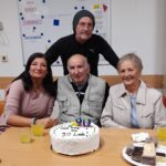 Štirje odrasli stojijo in sedijo za mizo s torto za 90. rojstni dan; v ospredju je slavljenec, ob njem pa trije spremljevalci, na mizi so pijača in pecivo.