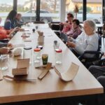 Starejše osebe sedijo ob dolgi mizi in iz kartona ter volne izdelujejo božično dekoracijo; pred njimi so kozarci, volnene niti in orodje za ustvarjanje.