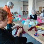 Skupina stanovalcev in animatorka ustvarjajo pisane božične okraske iz volne; na mizi so klobčiči volne, škarje, lepilo in delno izdelani okraski.