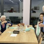 Starejši moški in ženske sedijo za mizo ter izdelujejo volnene božične figure; eden od stanovalcev drži lesen obroč ovit z volno, drugi urejajo posamezne niti.