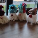 Vrsta končanih volnenih božičnih škratov v različnih barvah stoji na mizi; vsak ima volneno kapo, dolgo brado in okrasno kroglico kot nos.