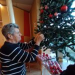 Stanovalec doma za starejše obeša rdeče okraske na božično jelko, ob njem je miza s prazničnimi kroglami.