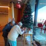 Tri stanovalke doma za starejše skupaj krasijo božično jelko ob oknu; v prostoru so mize, vedra in praznični okraski.