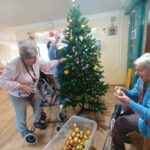 Stanovalke doma za starejše skupaj krasijo božično jelko z zlatimi okraski; ob drevesu je plastična posoda z okrasnimi kroglami.