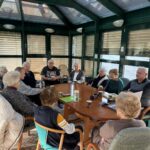 Skupina starejših udeležencev sedi okrog velike okrogle mize v svetlem zimskem vrtu ter sodeluje v literarnem krožku; na mizi so skodelice, knjige in zapiski.