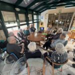 Starejši udeleženci sedijo v krogu okoli mize v zimskem vrtu, medtem ko poteka literarni krožek; v ozadju so steklene stene in vhod v prostor doma.