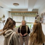 Starejša gospa v sobi doma starejših se pogovarja z dvema mladima obiskovalkama.