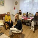 Starejša stanovalka sedi v sobi doma starejših, ob njej so tri mlade obiskovalke, ena bere knjigo.