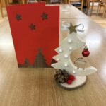 Na mizi sta praznična voščilnica in lesena dekoracija v obliki smrečice z božičnimi okraski.