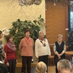 Skupina starejših žensk stoji v prostoru doma in poje ob prazničnem dogodku Miklavža.