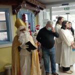 Miklavž v zlati opravi stoji v hodniku doma, ob njem skupina mladih v belem, eden izmed obiskovalcev nosi zaščitno masko.