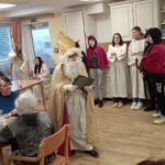 Miklavž in njegova spremljevalna skupina angelov in parkeljnov obiskujejo stanovalce v jedilnici doma, kjer razdeljujejo darila.