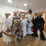 Skupina oseb v kostumih Miklavža, angelov in parkeljnov stoji pred okrašeno božično smrečico v notranjem prostoru doma.