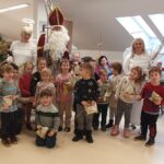 Skupina otrok z vzgojiteljicama ter obiskovalci v kostumih Miklavža in angelov pozira za fotografijo; otroci v rokah držijo darilca in šibe.