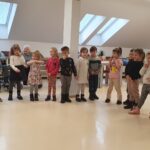 Otroci iz vrtca stojijo v vrsti in nastopajo pred stanovalci doma; ob strani kleči vzgojiteljica, v ozadju sedijo starejši stanovalci.