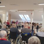 Otroci nastopajo v skupnem prostoru doma, okoli njih v polkrogu sedijo stanovalci na stolih in vozičkih; v ozadju je božično drevo.