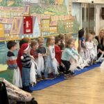 Skupina predšolskih otrok stoji v vrsti na modri podlagi in nastopa pred stanovalci doma za starejše. Otroci pozorno sledijo vzgojiteljicama, ki klečita ob njih. V ozadju je barvita stenska poslikava mesteca.