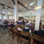 Velika skupina stanovalcev doma za starejše sedi v jedilnici oziroma skupnem prostoru in opazuje otroški nastop na sprednjem delu prostora. Med gledalci so tudi osebe na invalidskih vozičkih.