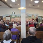 Številni stanovalci sedijo obrnjeni proti odru, kjer otroci v božičnih kapicah nastopajo. V ozadju je velika stenska poslikava in ura nad osrednjim delom prostora.