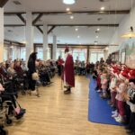 Dobro obiskan nastop v večjem prostoru doma za starejše, kjer otroci v božičnih kapicah stojijo na desni strani, na levi pa sedijo stanovalci. V sredini stoji Miklavž, ob njem pa spremljevalec v črnem kostumu.