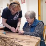 Starejša gospa pomaga drugi uporabnici pri vezanju zlatega traku na leseno vejo med ustvarjalno delavnico.