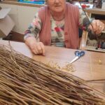 Starejša ženska sedi za mizo in drži dolgo vejo, ob njej pa leži kup tankih vejic in škarje.