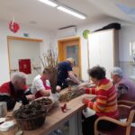 Skupina stanovalcev sedi za mizo v ustvarjalnici, kjer na dolgo vejo pritrjujejo zlate trakove, na mizi pa so tudi storži in orodje.