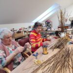 Dve starejši gospe sedita za mizo in navezujeta zlate trakove na veje, pred njima je velika zbirka tankih vejic in kolut zlatih trakov.