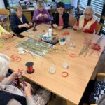 Skupina starejših žensk sedi okoli mize in na tanke vejice pritrjuje rdeče trakove za Miklavževe šibe.