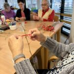 Bližnji pogled na roke starejše ženske, ki na tanko vejico zavezuje rdeč trak, v ozadju sedijo druge udeleženke delavnice.