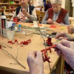 Roke stanovalke zavezujejo rdeč satenast trak na vejico, za mizo pa druge starejše ženske izdelujejo Miklavževe šibe.