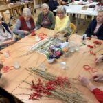 Večja skupina starejših žensk sedi okoli mize, pred njimi so kupi vejic in rdečih trakov, medtem ko skupaj izdelujejo Miklavževe šibe.