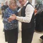 Moški folklorni plesalec v klobuku pleše z nasmejano starejšo gospo; v ozadju so drugi stanovalci in izvajalci.