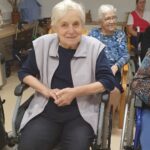 Starejša gospa na invalidskem vozičku se nasmehne proti kameri, v ozadju so drugi stanovalci.