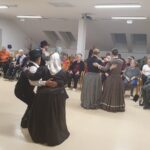 Več parov v folklornih oblačilih pleše pred občinstvom, ki sedi na stolih in invalidskih vozičkih.