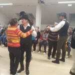 Folklorni plesalci in stanovalci plešejo v paru; nekateri gledalci opazujejo dogajanje iz ozadja.