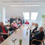 Skupina stanovalcev sedi za mizo v skupnem prostoru, ob njih stojijo birmanci in odrasli spremljevalec; na mizi so skodelice in drobni predmeti.