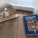 Na mizi so razstavljeni leseni božični okrasek z motivom jaslic, zvitki in knjižica z naslovom »Družinska pratika 2026«.